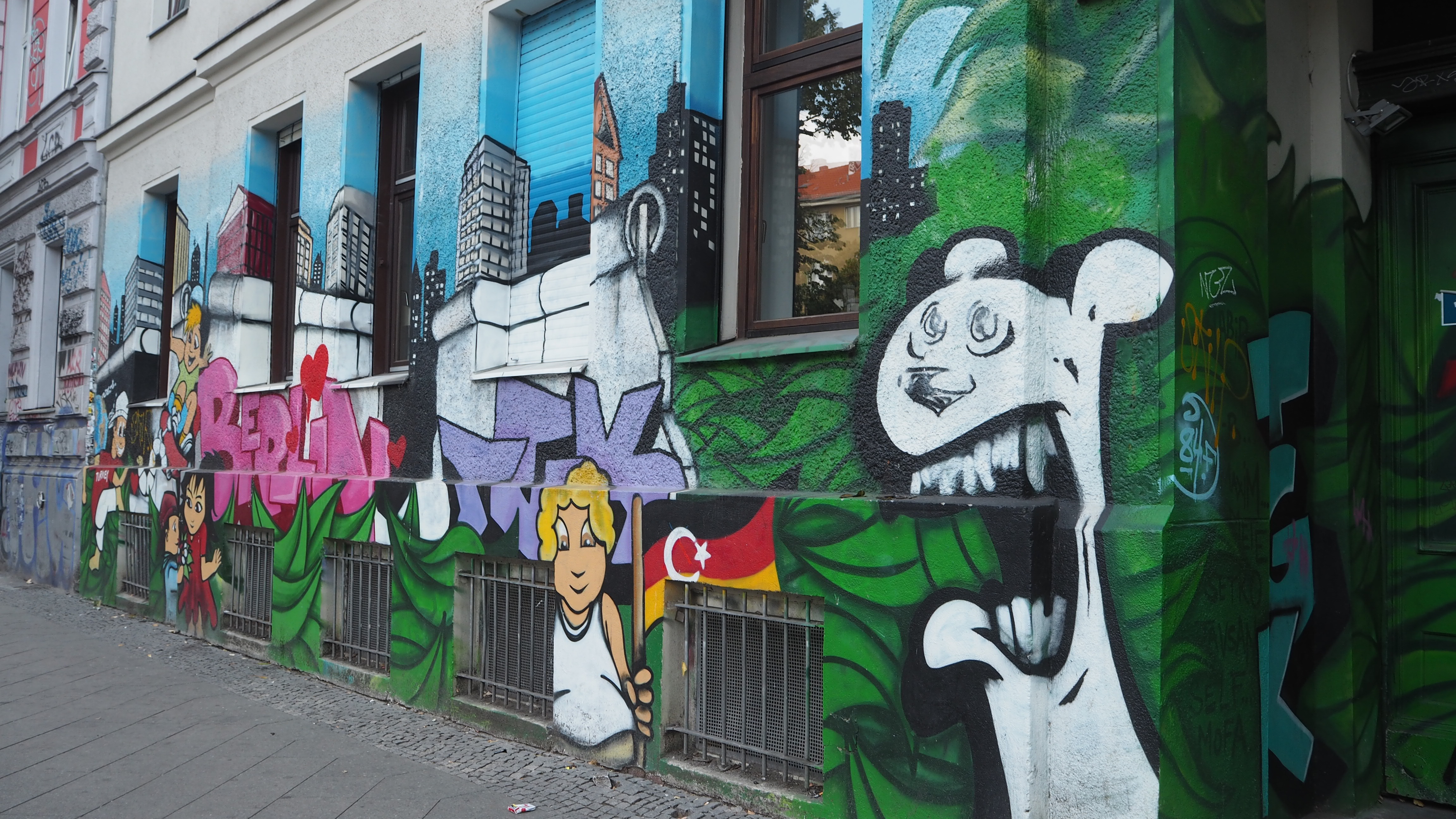 Street art on the wall of a building, with a cityscape and then a white Berlin bear, a child holding the German flag and the word Berlin