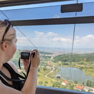 a girl with blonde hair taking a photo out of the window of a cable car on the Dajti Express. The view is of mountains and a lake